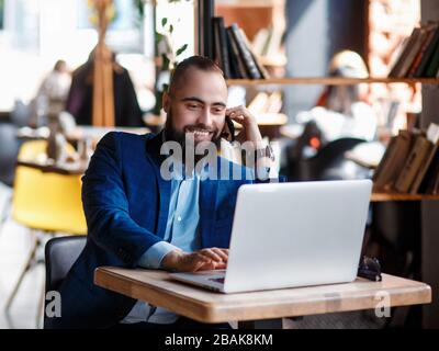 jeune homme d'affaires barbu au travail au bureau. Un homme est assis sur un ordinateur portable qui parle par téléphone mobile. Conversations téléphoniques dans un collègue Banque D'Images