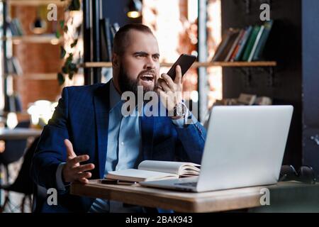 Un homme d'affaires barbu sérieux mène des conversations téléphoniques sur un téléphone mobile à l'ordinateur. Un homme mécontent fait peur au téléphone. Des citations de rage dans Banque D'Images