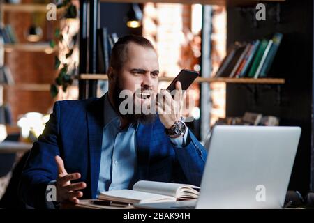 Un homme d'affaires barbu sérieux mène des conversations téléphoniques sur un téléphone mobile à l'ordinateur. Un homme mécontent fait peur au téléphone. Des citations de rage dans Banque D'Images