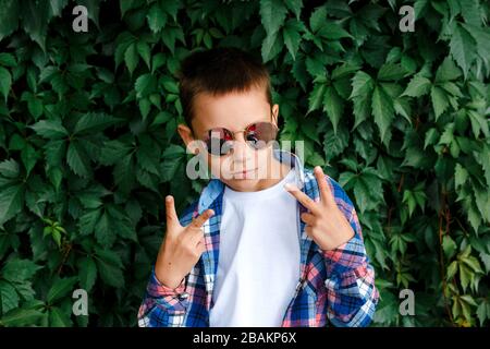 Un petit garçon élégant dans des lunettes de soleil rouges montre le signe de paix extérieur Banque D'Images