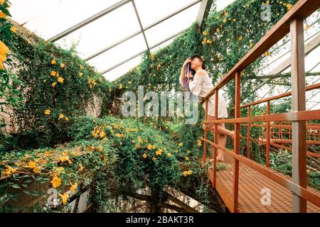 une jolie fille solitaire avec des readlocks multicolores dans une robe blanche se tient sur un escalier rouge dans une arche de fleurs. Serre avec tropical exotique p Banque D'Images