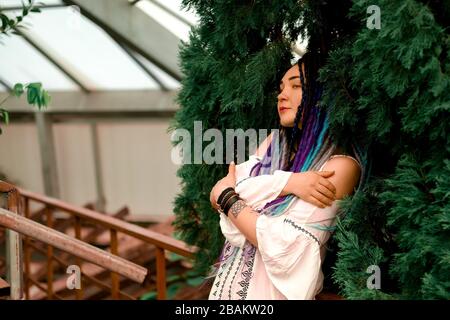 une jolie fille solitaire avec des dreadlocks multicolores dans une robe blanche est debout sur un escalier rouge près, cyprès vert, arbre de thuja. Serre avec ex Banque D'Images