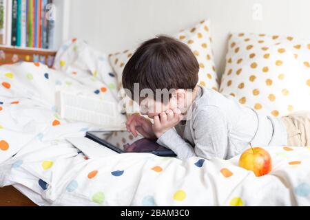 School Boy sur lit à la maison avec tablette numérique à la main, faire des devoirs. Formation à distance en ligne. Quarantaine Banque D'Images