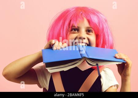 Fille de l'école avec vue heureuse isolée sur fond rose. Retour au concept d'école et d'éducation. Élève en uniforme scolaire avec perruque rose. Fille morde grand livre bleu. Banque D'Images