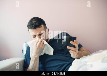 Jeune homme malade avec hanky. Le gars se trouve dans le lit avec une couverture éternuements et couvre son nez avec une serviette. Dans l'autre main tient une tasse. Isolé sur Banque D'Images