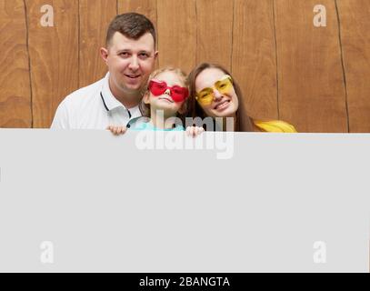 Famille souriante derrière un panneau blanc blanc sur fond de bois. Visages bizarres. Sortez de derrière une bannière, videz l'espace pour le sommet Banque D'Images