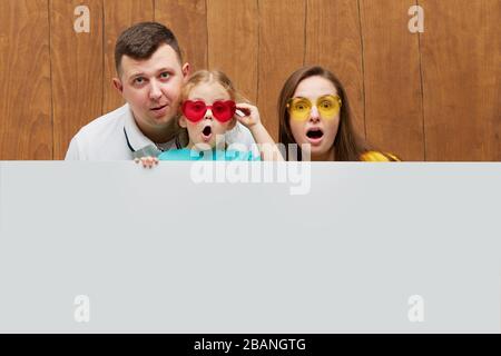 Famille souriante derrière un panneau blanc blanc sur fond de bois. Visages bizarres. Sortez de derrière une bannière, videz l'espace pour le sommet Banque D'Images