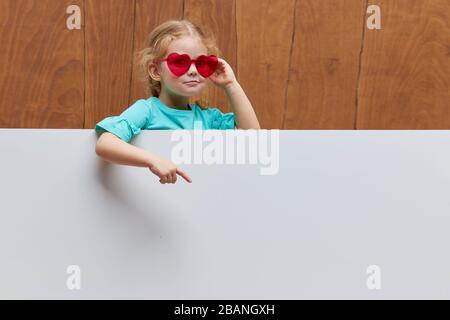 Petite fille souriante lunettes en forme de coeur debout derrière un panneau blanc blanc blanc contre fond en bois. Visage drôle. Sortez de derrière un ba Banque D'Images