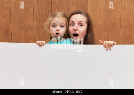 Famille souriante derrière un panneau blanc blanc sur fond de bois. Visages bizarres. Sortez de derrière une bannière, videz l'espace pour le sommet Banque D'Images