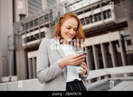 Une femme d'affaires qui a réussi à lire un courrier de confirmation sur son smartphone tout en tenant une tasse à café jetable en extérieur Banque D'Images