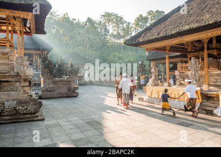 Famille balinaise dans une robe traditionnelle marchant sur la cour du temple en début de matinée misty lumière avec des frères tenant les mains jetant de longues ombres. Banque D'Images