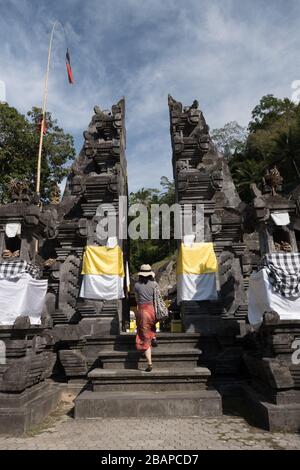 Femme vêtue de paille, sarong rouge et noir et haut noir marchant vers le haut des marches d'entrée du temple en pierre sculpté en début de matinée avec le ciel bleu. Banque D'Images