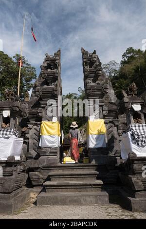 Femme vêtue de paille, sarong rouge et noir et haut noir marchant vers le haut des marches d'entrée du temple en pierre sculpté en début de matinée avec le ciel bleu. Banque D'Images