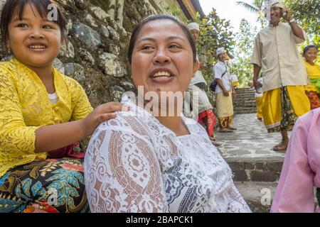 Mère balinaise portant un haut en dentelle blanche, souriante, jeune fille reposant main sur son épaule, assise sur le mur du temple avec Père en arrière-plan. Banque D'Images