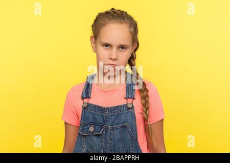 Portrait d'une petite fille mécontente et drôle et frustrée, avec une tresse dans des salopettes en denim, regardant un appareil photo avec une expression de visage mécontent et comique. Intérieur Banque D'Images