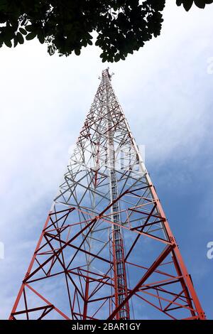 Cette image représente une grande tour de télécommunications debout à l'extérieur contre un ciel bleu clair. Il y a quelques nuages tordus et un petit tre vert Banque D'Images