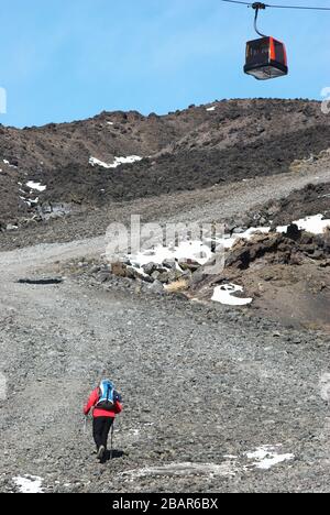 Un homme avec des bâtons de randonnée monte sur la pente raide et pierreuse jusqu'aux cratères du sommet de l'Etna, au-dessus d'une cabine de téléphérique de l'Etna Park, Sicile Banque D'Images