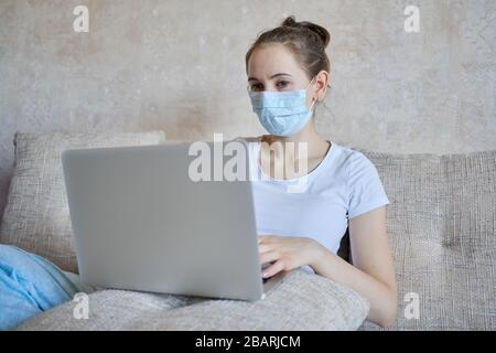 Une fille dans un masque médical est en quarantaine à la maison travaillant sur un ordinateur portable. Gros plan. Banque D'Images