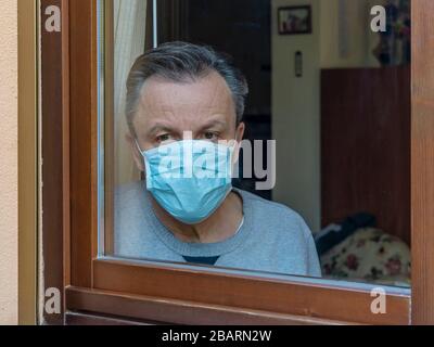 Homme italien avec masque de protection, forcé de rester à la maison en raison du coronavirus covid-19, disconsolate regarde la fenêtre de la maison Banque D'Images