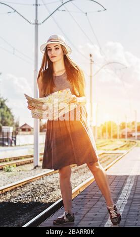 Recherche de son transport pour un nouveau voyage. Pleine longueur de belle et jeune femme en robe et chapeau en gardant la carte dans les mains et en regardant loin pendant stan Banque D'Images