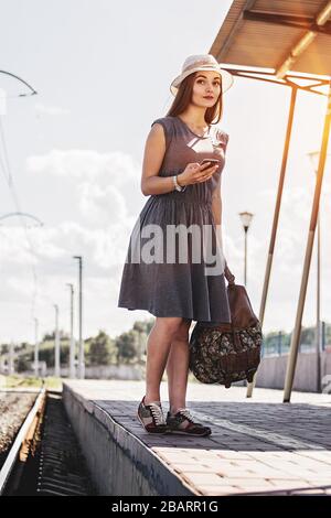 En attente de son transport pour commencer un nouveau voyage. Toute la longueur de la jeune femme séduisante en robe en gardant le téléphone dans une main et sac à dos dans une autre pendant Banque D'Images