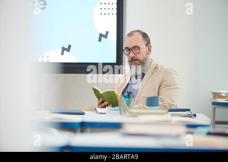 Portrait d'un professeur barbu senior assis au bureau dans la salle de classe scolaire et livre de lecture, espace de copie Banque D'Images