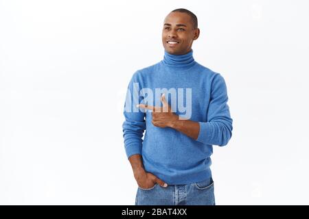 Jeune homme afro-américain réussi, vêtu d'un col de cygne bleu, regarde avec satisfaction le visage et pointe vers la gauche, souriant heureux de voir de bonnes occasions ou Banque D'Images