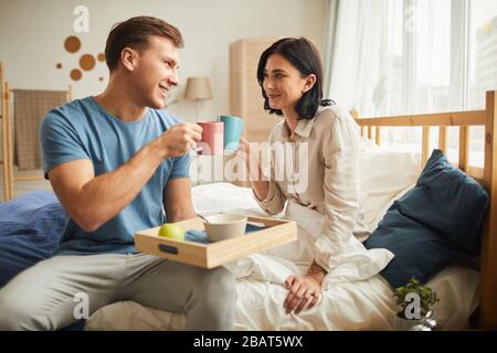 Vue latérale portrait de heureux jeune couple prenant le petit déjeuner dans le lit et clinking café tasses tout en regardant l'un l'autre avec amour, espace de copie Banque D'Images