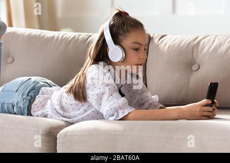 Adorable petite écolière posée sur un canapé, portant un casque sans fil moderne. Banque D'Images