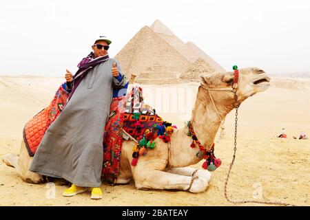 Un chauffeur de chameau pose à dos de chameau sur le plateau de Giza au Caire, en Egypte. Les grandes pyramides de Gizeh sont en arrière-plan. Banque D'Images