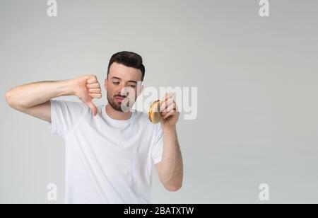 Jeune homme isolé en arrière-plan. Guy tient grand pouce vers le bas pour un hamburger insavoureux dans sa main. Cheeseburger malsain et gras. Homme barbu en chemise blanche Banque D'Images