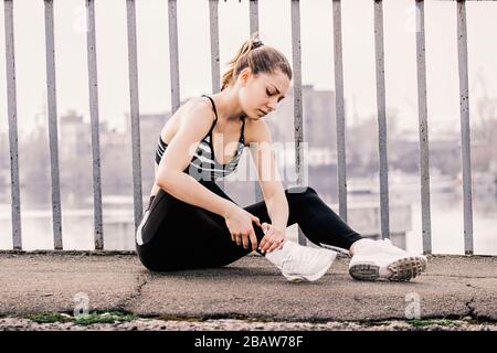 Jeune femme de fitness déçue dans des vêtements de sport élégants assis sur la route et tenant sa cheville de braconnage. Belle fille de sport triste ayant la douleur dans sa jambe Banque D'Images