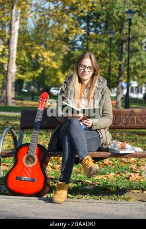 Jeune fille mignonne avec des lunettes lisant un livre sur le banc dans un parc, avec de la guitare acoustique rouge penchant contre le banc Banque D'Images