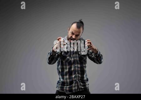 Homme barbu aux cheveux noirs peignés, portant une chemise à carreaux, montrant une grimace agressive et des gestes d'écrasement avec ses mains vers le haut. Port avant demi-longueur Banque D'Images