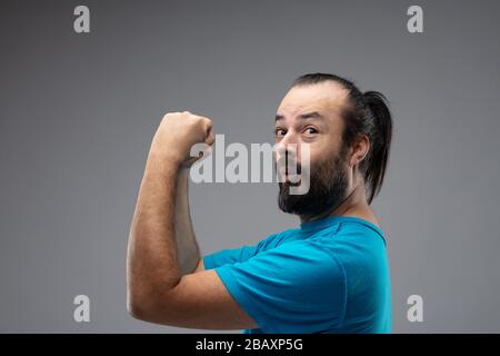 Homme barbu au look heureux dans un t-shirt bleu et avec de longs cheveux noirs peignés, exprimant le geste gagnant avec ses mains vers le haut et les poings serrés. Gros plan s Banque D'Images