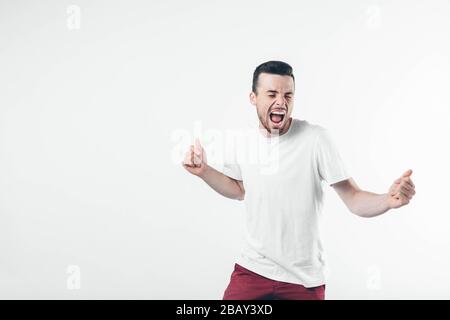 Jeune homme brunette avec barbe vêtu d'un T-shirt blanc. Guy dansant et chantant avec une large bouche ouverte et des yeux fermés. Isolé sur fond clair. Banque D'Images