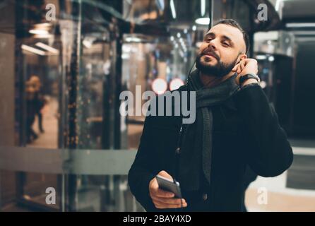 Vue latérale du jeune homme barbu, vêtu de vêtements incasuels, il se tient dans la rue et utilise un smartphone. Banque D'Images