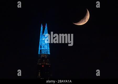 Les 20 premiers articles du gratte-ciel Shard sont illuminés en bleu NHS dans le cadre d'un hommage national qui remercie tout le personnel des services de santé et les travailleurs clés qui luttent actuellement contre la propagation du virus COVID-19. Une lune de Crescent qui se couche sur le Shard juste après minuit. Londres, Royaume-Uni. Banque D'Images