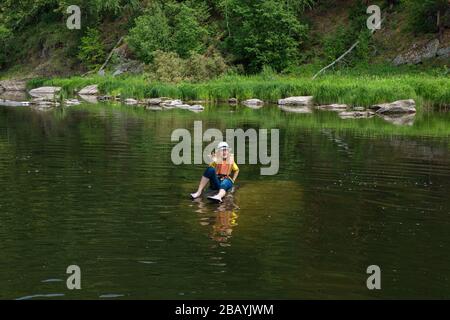 Criant jeune fille dans chapeau, short de Jean et gilet de vie orange se trouve sur une pierre plate au milieu du lac forestier qui l'étend la main vers l'avant. Sauvez-moi. BO Banque D'Images