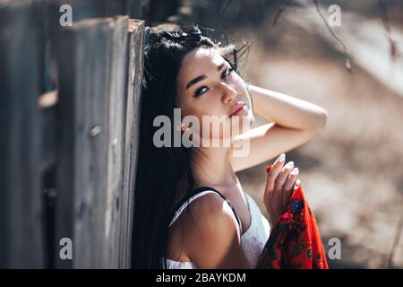 Portrait d'une belle fille à cheveux noirs dans une robe blanche vintage debout près de la clôture en bois.modèle de jeune femme posant dans un style national russe . rouge Banque D'Images