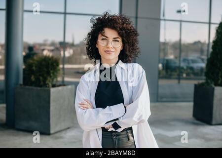 Une charmante femme caucasienne aux cheveux et aux verres raillés se pose avec des mains croisées et regarde l'appareil photo Banque D'Images