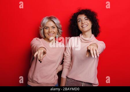Une femme brunette aux cheveux bouclés et son ami blond pointant vers l'appareil photo tout en posant sur un fond rouge Banque D'Images