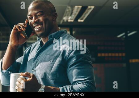 Un jeune homme souriant avec une tasse de café en main parlant sur le téléphone portable dans un bureau moderne. Un homme d'affaires heureux qui appelle au bureau. Banque D'Images