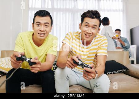 Les gars vietnamiens excités jouant à des jeux vidéo à la maison, leurs amis parlent en arrière-plan Banque D'Images