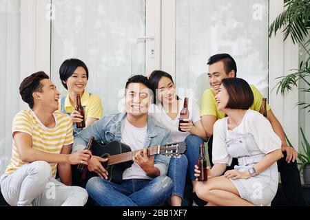 Joyeux beau jeune homme vietnamien jouant de la guitare pour ses amis buvant et parler à la fête de la maison Banque D'Images