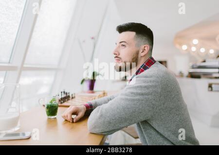 Jeune homme barbu dans un café. Il est assis à une longue table près de la fenêtre. Boisson avec des feuilles vertes à imprimé smartphone sur le bureau. Homme pensant et regardant dehors Banque D'Images