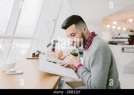 Jeune homme barbu dans un café. Il est assis à une longue table près de la fenêtre. Guy tient le verre avec des feuilles vertes d'une main. Un autre sur le magazine que l'homme lit Banque D'Images