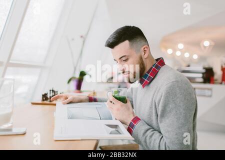 Jeune homme barbu dans un café. Il est assis à une longue table près de la fenêtre. Guy tient le verre avec des feuilles vertes d'une main. Un autre sur le magazine que l'homme regardant Banque D'Images