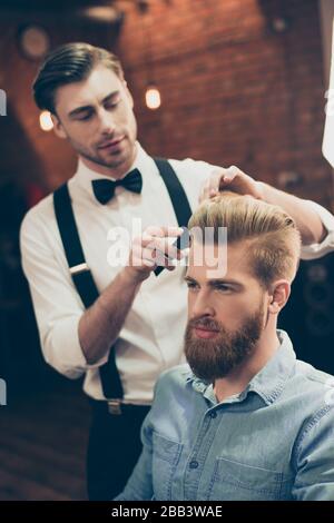 Coiffeur chic habillée beau styliste fait une coiffure parfaite à un gars barbu dans un Jean caual tenue. Concentré et sérieux Banque D'Images