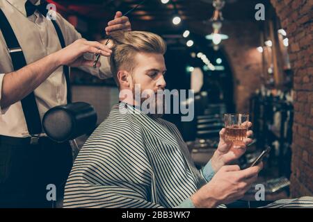 Détendez-vous au salon de coiffure. Vue latérale de l'homme barbu rouge beau buvant scotch et de naviguer sur son pda, tout en obtenant une coupe de cheveux Banque D'Images